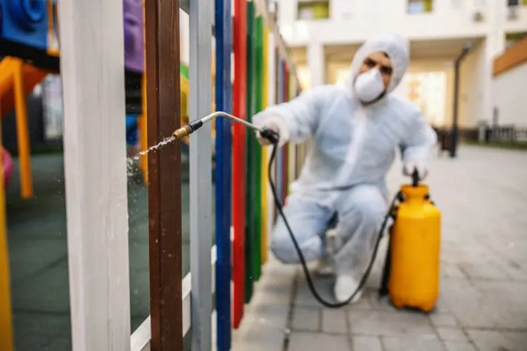 Profissional em traje de proteção faz limpeza escolar externa com pulverizador para garantir segurança.