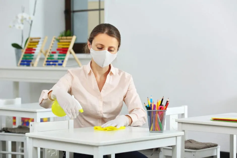 Funcionária com máscara e luvas higieniza mesa de sala de aula para manter limpeza escolar.