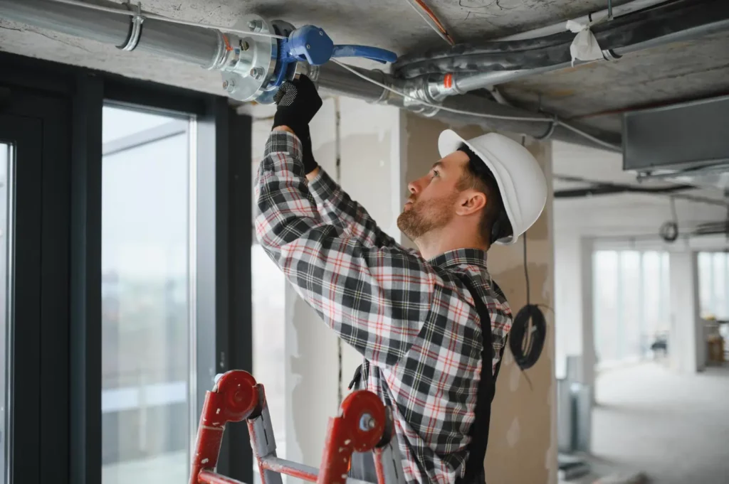 Supervisor, auxiliar, técnico e oficial de manutenção predial: qual a diferença? Profissional de manutencao predial com capacete ajustando tubulação no teto de edifício em obra
