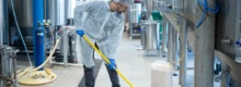 Profissional de limpeza técnica com uniforme branco e touca higiênica esfregando o piso de uma fábrica cercada por tanques de aço inoxidável.