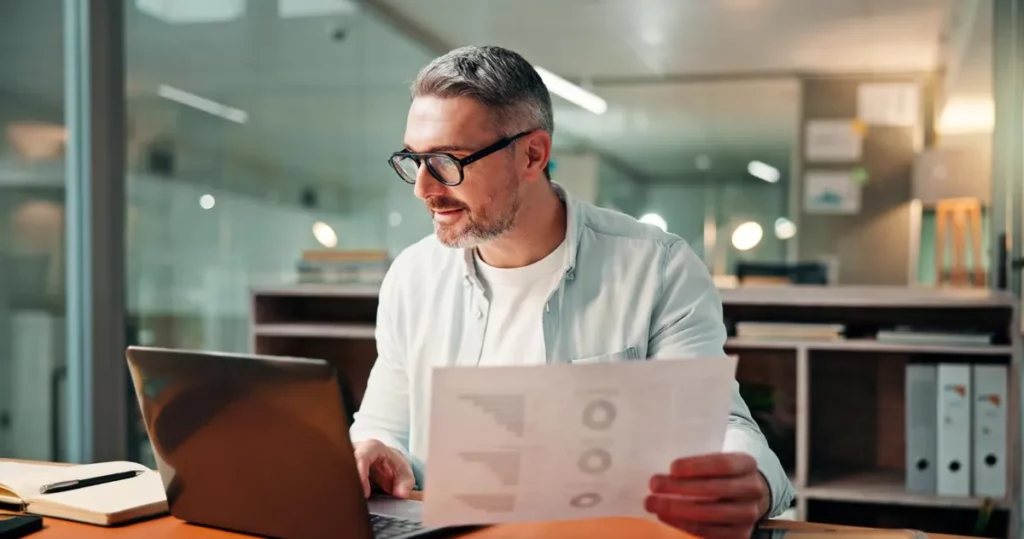 Profissional de escritório analisando gráficos em relatório enquanto trabalha no notebook, relacionado à NR-1 e saúde mental